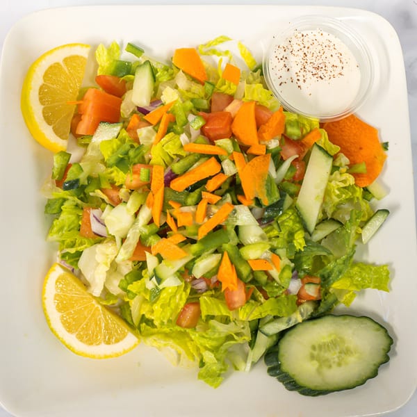 A fresh side salad with lettuce, carrots, cucumbers, tomatoes, and a side of dressing.